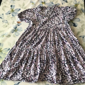 Blommig klänning  - Charmig blommig klänning i en mix av svart, vit och rosa. Klänningen har korta ärmar och en v-ringad hals. Perfekt för en somrig look med sitt luftiga och bekväma material.