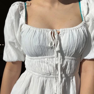 Vit klänning med puffärmar - Säljer en söt vit klänning med puffiga kortärmar och knytning framtill. Klänningen har en smickrande passform med resår i midjan och en luftig känsla. Perfekt för varma dagar!