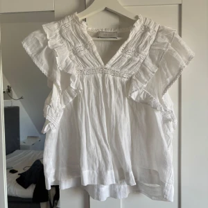 Blus  - Säljer en söt vit blus från Neo Noir med volangdetaljer och korta ärmar. Blusen har en luftig passform och är perfekt för varma dagar. Den har en vacker struktur och är tillverkad i ett lätt material.