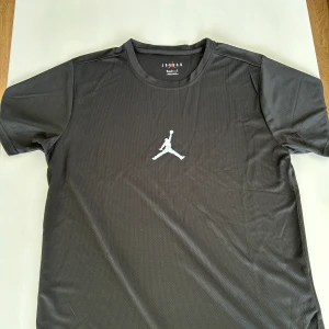 Svart kortärmad t-shirt från Jordan - Säljer en svart kortärmad t-shirt från Jordan med det ikoniska logotypen på bröstet. Tillverkad i ett lätt och andningsbart syntetmaterial, perfekt för träning eller vardagsbruk. T-shirten har en klassisk rund halsringning och en stilren design. Storlek XL men passar som L