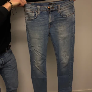 Blå jeans från Nudie Jeans - Säljer ett par snygga blå jeans från Nudie Jeans. De har en klassisk femficksdesign med orange sömmar och knappgylf. Perfekta för en avslappnad stil.