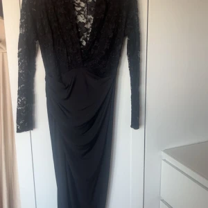 Svart långklänning med spetsdetaljer och slits - Elegant svart långklänning med v-ringad spetsöverdel och långa ärmar. Klänningen har en figurnära passform och är perfekt för festliga tillfällen. Den vackra spetsen ger en sofistikerad touch. Använd ett tillfälle. Hund finns i hemmet.