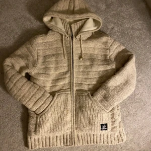 Beige stickad Jacka/Cardigan -  Stickad jacka/Cardigan från Shakaloha med huva och dragkedja. Perfekt nu när våren närmar sig! Finns det frågor är det bara att höra av sig😁
