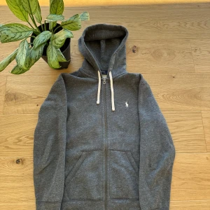 Grå hoodie från Polo Ralph Lauren - Säljer en stilren grå hoodie från Polo Ralph Lauren med dragkedja och broderad logga på bröstet. Tröjan har en justerbar huva med vita snören och är perfekt för en avslappnad look.