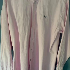 Ljusrosa skjorta från Fred Perry - Snygg ljusrosa skjorta från Fred Perry med klassisk krage och knappar framtill. Skjortan har långa ärmar och en broderad logotyp på bröstet. Perfekt för en stilren look.