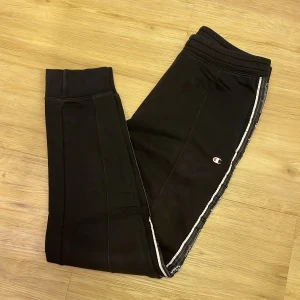 Svarta mjukisbyxor från Champion - Säljer ett par svarta mjukisbyxor från Champion med vita detaljer längs sidorna. Byxorna har en elastisk midja och är tillverkade i en mjuk bomullsblandning för extra komfort. Perfekta för en avslappnad stil.