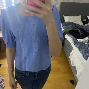 Blå blus med puffärmar - Säljer en söt blå blus med puffärmar och vågigt hålmönster. Perfekt för en stilren och chic look. Blusen har en rund hals och korta ärmar som ger en feminin touch.