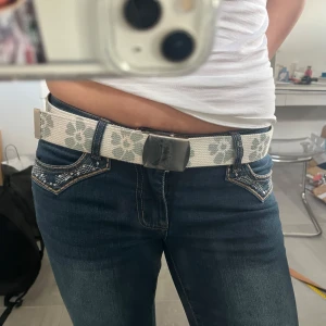 Vitt bälte med blommönster - Snyggt vitt bälte med grått blommönster och ett stilrent metallspänne. Perfekt för att ge en extra touch till din outfit. Passar bra till jeans och andra casual kläder. 100 cm långt. 