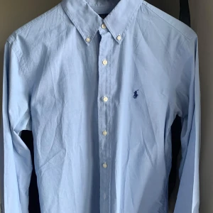 Ralph Lauren skjorta - Snygg ljusblå skjorta från Ralph Lauren med klassisk krage och broderad logga på bröstet. Skjortan har långa ärmar och knappar framtill. Perfekt för en stilren look.
