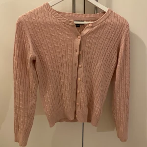 Rosa stickad kofta med knappar - Säljer en söt rosa stickad kofta med ett klassiskt flätmönster. Koftan har långa ärmar och knäpps med matchande knappar framtill. Säljer bara för att den blivit för liten💗