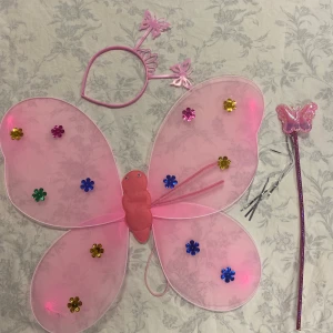 Fjärilsvingen med diadem och trollstav - Säljer ett färgglatt fjärilsset som inkluderar vingar, diadem och trollstav. Finns i rosa, lila och gul med glittriga detaljer och blommor. Perfekt för lek och utklädnad!  75kr/paket/3delar , helt ny,  skicka med postnord 