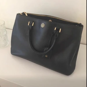 Tory Burch Robinson Double zip tote med kvitto - Snygg svart handväska från Tory Burch i skinn med guldfärgade detaljer. Väskan har en avtagbar axelrem och dragkedja upptill. Insidan är fodrad med ett mönster av logotypen. Perfekt för att bära dina dagliga nödvändigheter med stil. Inkl kvitto(5300kr) och dustbag.