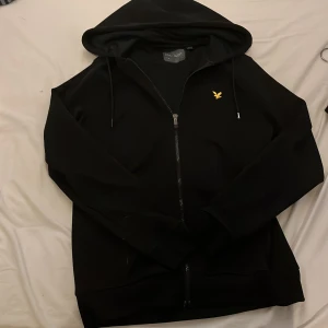 Svart hoodie från Lyle & Scott - Säljer en stilren svart hoodie från Lyle & Scott med dragkedja och en liten gul logga på bröstet. Perfekt för en avslappnad look. Den har en praktisk huva med snören.