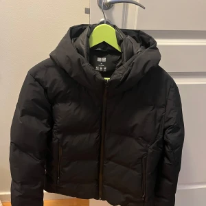 Svart pufferjacka från Uniqlo - Säljer en svart pufferjacka från Uniqlo. Jackan har en dragkedja framtill och en huva för extra skydd. Perfekt för kyligare dagar med sin lätta och vattentäta design. Jättefint skicka! 