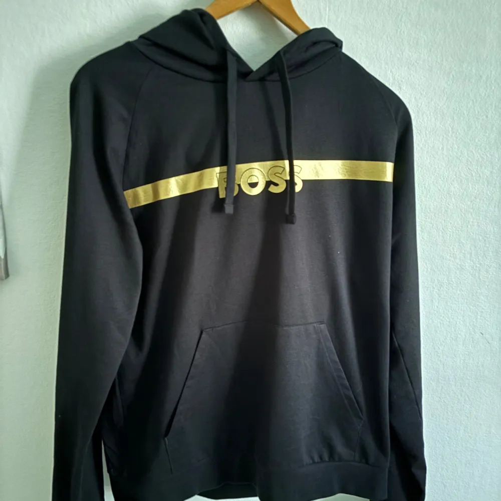 Snygg svart hoodie från Boss med en gyllene rand och texten 'BOSS' på bröstet. Perfekt för en stilren och avslappnad look. Den har en klassisk känguruficka och justerbar huva med dragsko.. Neuletakit & Villapaidat.