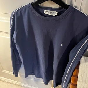 Mörkblå långärmad tröja från Yves Saint Laurent - Säljer en stilren mörkblå långärmad tröja från Yves Saint Laurent med vita ränder längs ärmarna och en liten logga på bröstet. Perfekt för en avslappnad men ändå elegant look. Passar bra till både jeans och chinos.