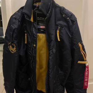 Svart bomberjacka från Alpha Industries - Säljer en svart bomberjacka från Alpha Industries med gul insida och detaljer. Jackan har dragkedja och en ikonisk 'Remove Before Flight'-tag. Perfekt för en cool och avslappnad stil.