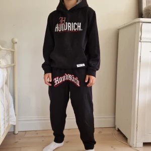 Hoodrich, byxor och hoodie - Hoodie: L, 12-13 Y. Byxor: M, 10-12 Y. Passar till dig som är 138-148 cm lång.  Svart hoodie från Hoodrich med stor vit och röd logga framtill och textdetaljer på ryggen. Tröjan har huva och långa ärmar. Mjuk och skön, perfekt för chill eller streetwear. Matchar med byxor i samma stil. Skön fleece på insidan. Pufftryck och screentryksgrafik.