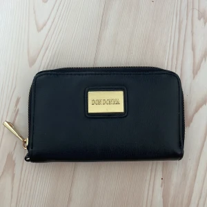 Svart plånbok från Don Donna - Snygg svart plånbok från Don Donna i skinnimitation med guldfärgade detaljer och dragkedja runtom. Flera kortfack, myntfack med dragkedja och plats för sedlar. Perfekt storlek för att ha med sig överallt och håller ordning på allt viktigt.