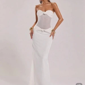 Vit axelbandslös långklänning - Supersnygg vit långklänning med axelbandslös design och figurnära passform. Klänningen har transparent mesh i midjan, knutdetaljer vid byst och höft samt en elegant, slät kjol. Perfekt för dig som vill sticka ut med en stilren och modern look. Endast provad på och säljs pga för liten storlek!