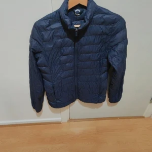 Mörkblå superlätt dunjacka XS - Säljer en mörkblå superlätt dunjacka från C i storlek XS. Jackan har quiltad design, hög krage och dragkedja framtill. Perfekt för kyliga dagar och enkel att bära med sig. Snygg och stilren modell som funkar till det mesta.