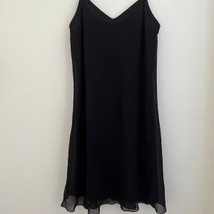 Svart kortklänning - En svart, enkel och flowy kortklänning med tunna axelband och v-ringning. Klänningen har en lätt, luftig känsla och faller snyggt med en något vågig nederkant. Perfekt för dig som gillar minimalistisk stil och vill ha något bekvämt och stilrent.