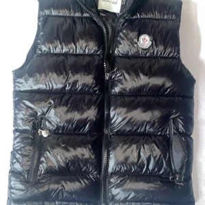 Svart dunväst från Moncler - Snygg svart dunväst från Moncler med glansig finish och klassisk logga på bröstet. Västen har två dragkedjeförsedda fickor och hög krage. Perfekt att slänga över en hoodie för en cool look. Riktigt bra passform och västen är i ny skick. 