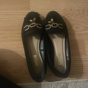 Svarta loafers med gulddetaljer Primark - Stilrena svarta loafers från Primark med snygga guldfärgade kedjedetaljer och tofs framtill. Skorna har platt sula och rund tå, tillverkade i mockaliknande material. Perfekta för dig som gillar klassisk men trendig stil.