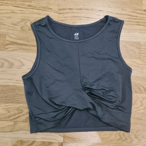 Mörkgrå sportig linnetopp från H&M - Mörkgrå, ärmlös topp från H&M Sport. Toppen har en croppad passform och en snygg twistad detalj framtill. Tillverkad i stretchigt material med 87% polyester och 13% elastan. Något stor i storleken, jag tycker mer det är en L.