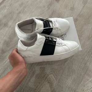 Valentino Garavani Open sneakers vit/svart - Snygga Valentino Garavani Open sneakers i vitt skinn med bred svart läderdetalj över vristen. Klassisk rund tå, vita snören och diskreta nitar på sulan. Perfekta för dig som gillar stilrena och lyxiga sneakers med en twist. Kvitto och allt finns med
