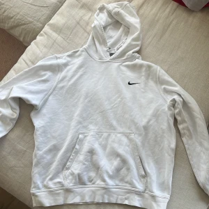 Vit hoodie från Nike med logga - Säljer en clean vit hoodie från Nike med klassisk svart swoosh broderad på bröstet. Tröjan har huva med snörning och en stor magficka. Perfekt för dig som gillar enkel och sportig stil.