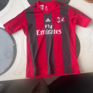 AC Milan Adidas fotbollströja röd/svart - AC Milan fotbollströja från Adidas i röd och svart med breda vertikala ränder. Tryckta loggor för Fly Emirates och klubbmärke på bröstet. Tillverkad i lätt och ventilerande Climalite-material, perfekt för träning eller match.