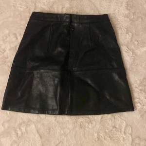 Svart skinnkjol från New Look Petite - Snygg svart kort skinnkjol från New Look Petite. Kjolen har en stilren design med markerade sömmar och hög midja. Perfekt för dig som gillar en edgy och trendig look. Passar till många olika outfits och är lätt att matcha.