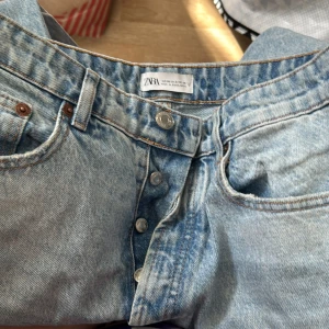 Ljusblå raka jeans från Zara - Säljer ett par ljusblå jeans från Zara med klassisk femficksmodell och knäppning med flera silverfärgade knappar. Jeansen har raka ben och är tillverkade i robust denim. Perfekta för en avslappnad och trendig look.