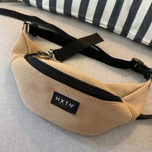 Beige magväska från HXTN Supply - Snygg beige magväska från HXTN Supply med svart justerbart band och dragkedja. Väskan är i mesh-liknande syntetmaterial och har en stilren svart logotyp framtill. Perfekt för att bära det viktigaste på stan eller festival.