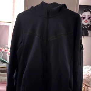 Mörkblå Nike Tech Zip Up - Mörkblå Nike Tech zip up hoodie med dragkedja, huva och fickor. Svart linje över bröstet och ärmficka med dragkedja. Använd några gånger men inga defekter förutom snöret på ärmfickan. S men rätt stor i storleken så hade nog passat M också!