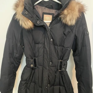 Svart dunjacka med äkta päls från Esprit - Snygg svart dunjacka från Esprit med quiltad design och stor huva med stor fluffig äkta päls. Jackan har bälte i midjan för en schysst siluett och stängs med både dragkedja och knappar. Perfekt för kalla dagar när du vill vara både varm och stilren. 