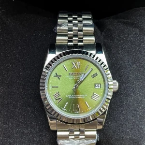 Seiko silverfärgad klocka med grön urtavla - Stilren Seiko klocka med grön urtavla och romerska siffror. Boetten och länken är i blankt silverfärgat stål med räfflad bezel. Klockan har datumvisning vid kl 3 och klassisk länk i metall. Perfekt accessoar för dig som vill sticka ut med en unik färg på urtavlan.