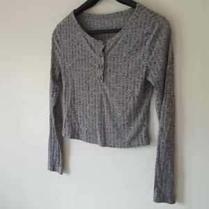 Grå ribbad långärmad topp med knappar strl S - Grå långärmad topp med ribbat mönster och fyra knappar framtill. Croppad passform som sitter snyggt till både jeans och kjol. Mjuk och stretchig i materialet, perfekt för lager-på-lager. Enkel men ändå trendig stil.  Helt & fint skick