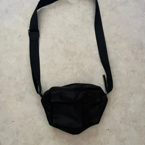 Svart axelväska med fickor - En svart axelväska i syntetmaterial med justerbart band. Väskan har två praktiska fickor framtill med lock och en dragkedja upptill. Perfekt storlek för att bära med sig det viktigaste och enkel att matcha till olika outfits.