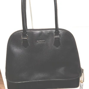 Svart handväska från Part Two - Stilren svart handväska från Part Two med rundad form och dubbla handtag. Väskan är tillverkad i pvc  med en lätt glansig finish och har en dragkedja upptill. Perfekt storlek för att få plats med allt du behöver till vardagen.