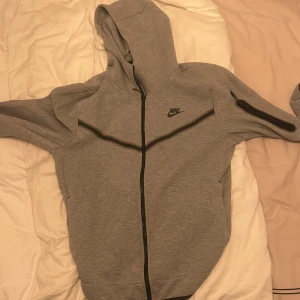 Grå hoodiejacka från Nike, M - Snygg grå hoodiejacka från Nike med hel dragkedja och svart logga på bröstet. Jackan har huva, svarta detaljer och en extra ficka med dragkedja på ena ärmen. Perfekt för chill eller träning, riktigt skön passform. 