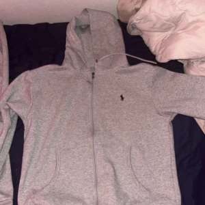 Grå hoodie från Polo Ralph Lauren - Snygg ljusgrå hoodie från Polo Ralph Lauren med dragkedja och klassisk logga på bröstet. Tröjan har huva med snörning, två fickor framtill och är tillverkad i mjuk bomullsmix. Perfekt för chill dagar eller när du vill ha en clean streetstyle.