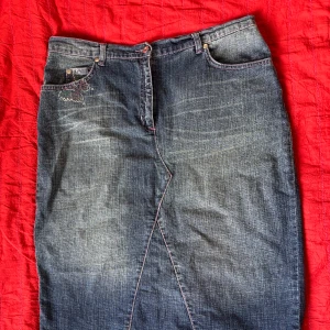 Jeanskjol med fjärilsdetalj och slits - Mörkblå längre jeanskjol med rosa sömmar och snygg slits bak. Fickor fram och bak en av bakfickorna  har en glittrig fjäril. 