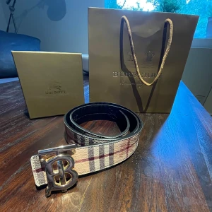 Burberry rutig skinnbälte med BB-spänne - Snyggt Burberry-bälte i klassiskt rutigt mönster med beige, svart och röd färg. Bältet har ett stort metallspänne med BB-logga och är gjort i äkta skinn. Perfekt accessoar för att lyfta din outfit med en lyxig touch. Pris kan diskuteras 
