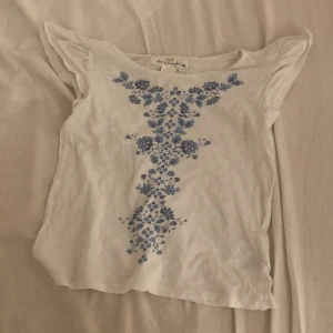 Vit blus med blå broderi från L.O.G.G. - Supersöt vit blus från L.O.G.G. med blått blommigt broderi framtill. Blusen har korta, volangiga ärmar och en lätt, luftig känsla. Perfekt för dig som gillar romantiska detaljer och vill ha något unikt i garderoben.