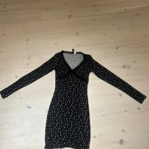 Svart blommig kortklänning med v-ringning - Supersöt svart kortklänning med små blommor, långärmad och v-ringad. Perfekt för dig som gillar en feminin och trendig stil. Klänningen har en smickrande passform och är lätt att styla med sneakers eller boots.