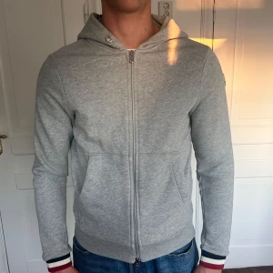 Grå Moncler hoodie med dragkedja - Snygg grå hoodie från Moncler med hel dragkedja, huva och Moncler-logga på ärmen. Tröjan har ribbade muddar med röd, vit och marinblå detalj samt fickor framtill. Snöret saknas på bilderna men skickas med vid köp. I bra skick☝️🪅pris kan diskuteras