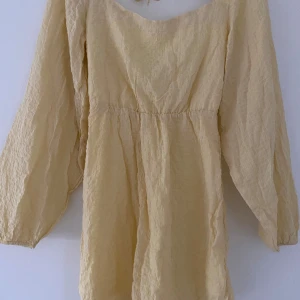 Ljusgul omlottklänning - Superfin ljusgul kortklänning med omlottdetalj och knytning i ryggen. Klänningen har långa, luftiga ärmar och är gjord i ett krinklat bomullstyg som känns lätt och somrigt. Perfekt för dig som gillar en avslappnad men söt stil.