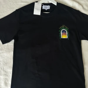 Svart t-shirt från Casablanca - Helt ny svart t-shirt från Casablanca med färgglad tryckt grafik av en byggnad och gröna blad både fram och bak. T-shirten har rund hals och korta ärmar. Perfekt för dig som gillar unika prints och streetwear vibes. Storlek S. 